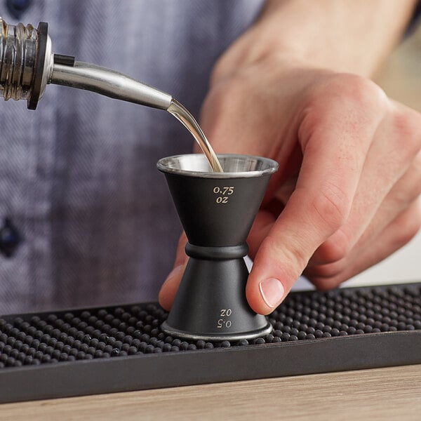 A close-up of a hand using a black Acopa Japanese jigger to pour liquid into a small glass.
