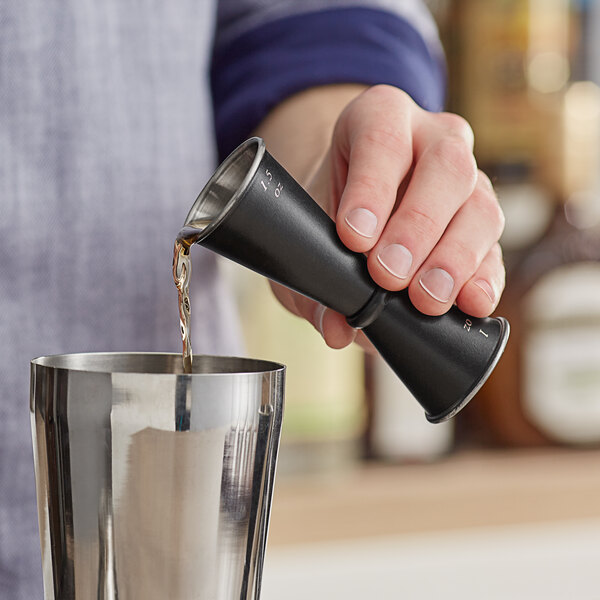 A hand holding a black Acopa Japanese Jigger pouring liquid into a metal cup.
