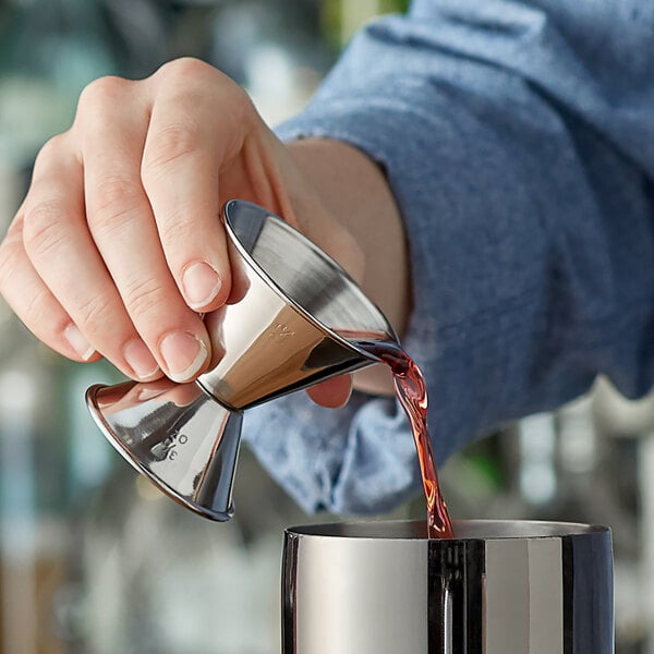 A person using a stainless steel classic jigger to pour liquid into a metal cocktail shaker.