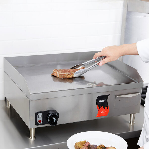 A chef using tongs to cook a steak on a Vollrath electric griddle on a counter.