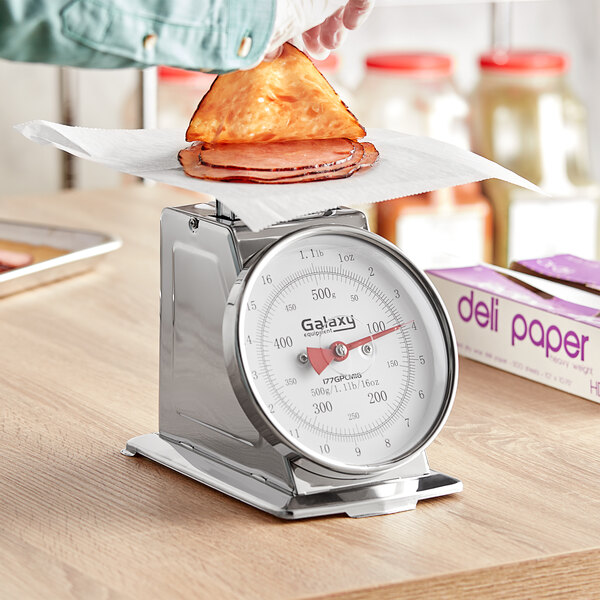 A person using a Galaxy mechanical portion scale to weigh a sandwich on a wood counter.