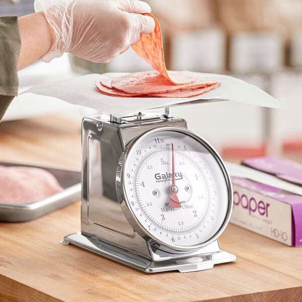A person using a Galaxy 10 lb. mechanical portion scale to weigh meat on a wooden surface.