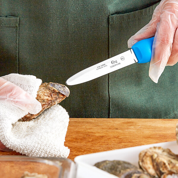 A person using a Choice 4" Galveston Style Oyster Knife with a blue handle to cut up oysters.