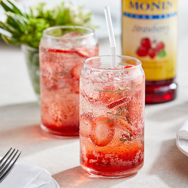 A glass of ice with a red drink flavored with Monin Sugar Free Strawberry Fruit Syrup, with a bottle of the syrup on the table.