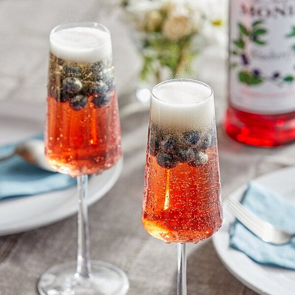 A glass of champagne with blueberries and a bottle of Monin blueberry syrup on a table.