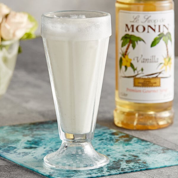A glass of milkshake on a blue mat next to a bottle of Monin Premium Vanilla Flavoring Syrup.