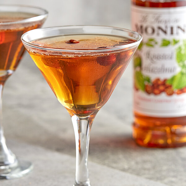 A glass of Monin roasted hazelnut flavored liquid on a table next to a bottle of Monin syrup.