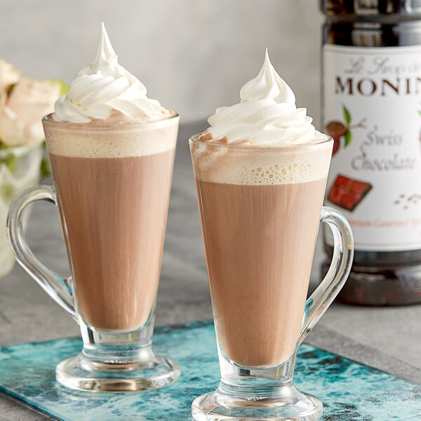 Two glasses of chocolate milk with whipped cream on a table with Monin Premium Swiss Chocolate Flavoring Syrup.
