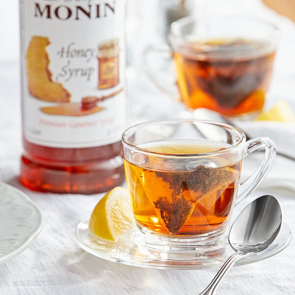 A glass cup of tea with Monin honey syrup on a saucer with a spoon and lemon slice.