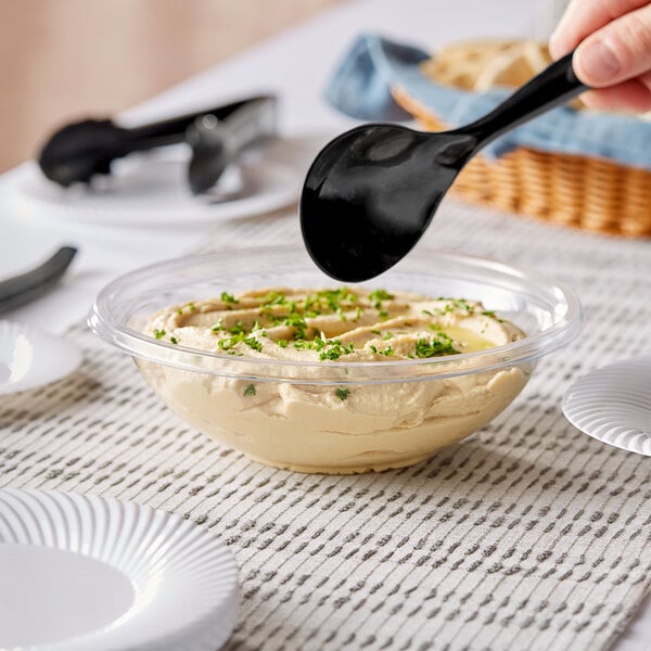 A person holding a black spoon over a bowl of hummus.