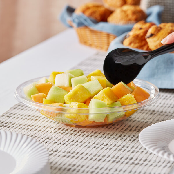 A Visions clear plastic bowl filled with fruit on a table with a spoon.