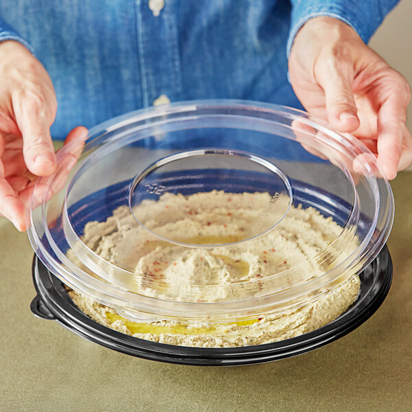 A person holding a Visions clear plastic lid over a bowl of hummus.