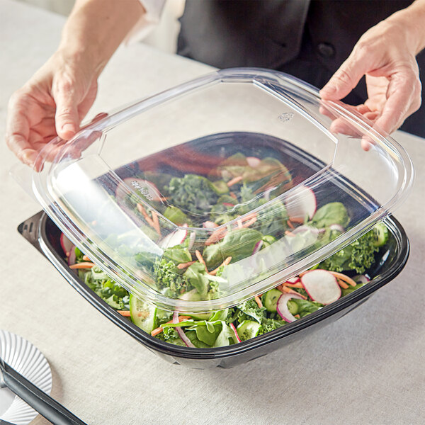 A person holding a Visions clear plastic dome lid over a salad in a clear plastic bowl.