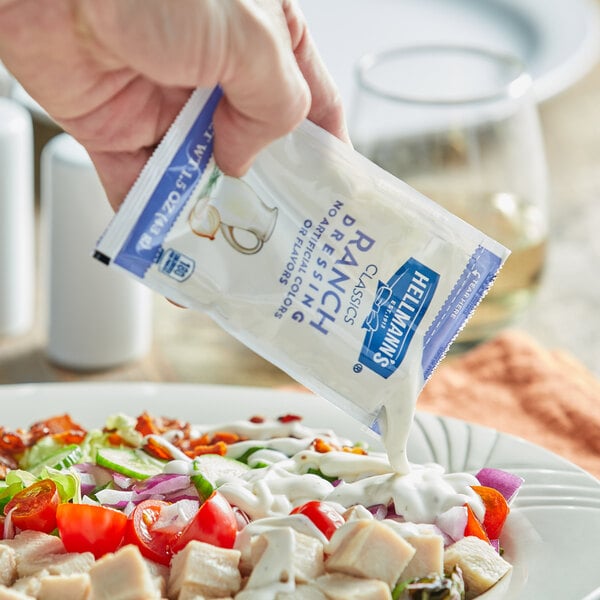 A hand pouring Hellmann's ranch dressing from a packet onto a salad.