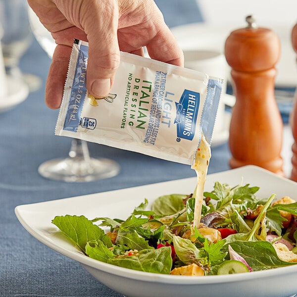 A hand pouring Hellmann's Light Italian Dressing from a small packet onto a salad.