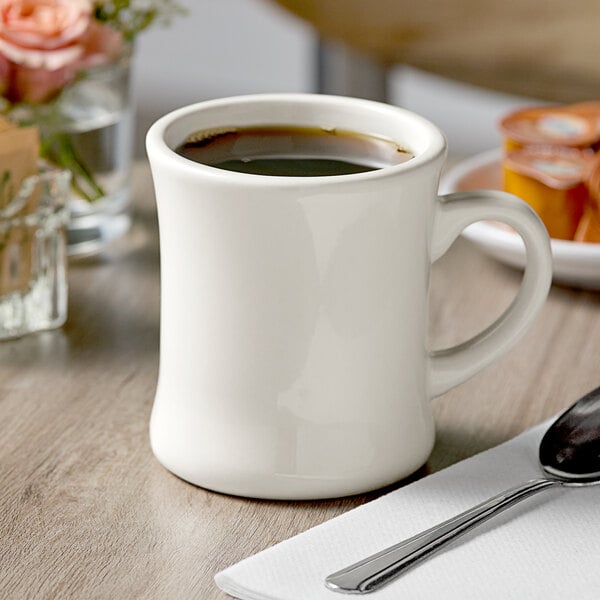 A white Acopa stoneware coffee mug with brown liquid and a spoon on a napkin.