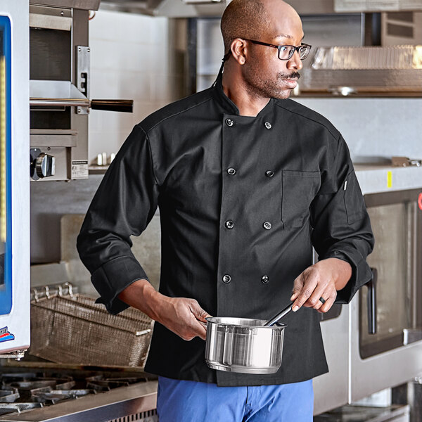 A man in a Uncommon Chef black long sleeve chef coat holding a pot.