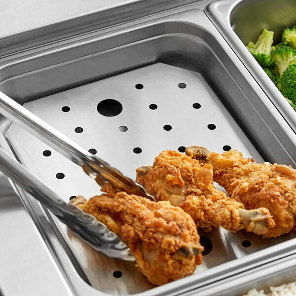 A stainless steel steam table pan with a false bottom on a counter with a tray of chicken and broccoli and tongs.