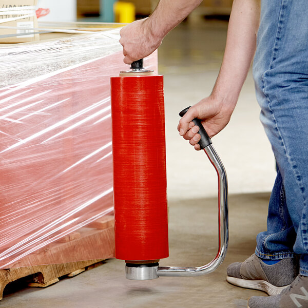 A person using a Lavex heavy-duty steel stretch wrap dispenser to wrap a box with red plastic.