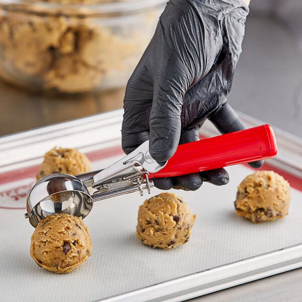 A person in a black glove using a red Choice 24 thumb press to scoop chocolate chip cookie dough.