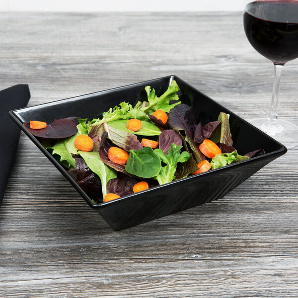 A black Siciliano square bowl filled with salad on a table with a glass of wine.