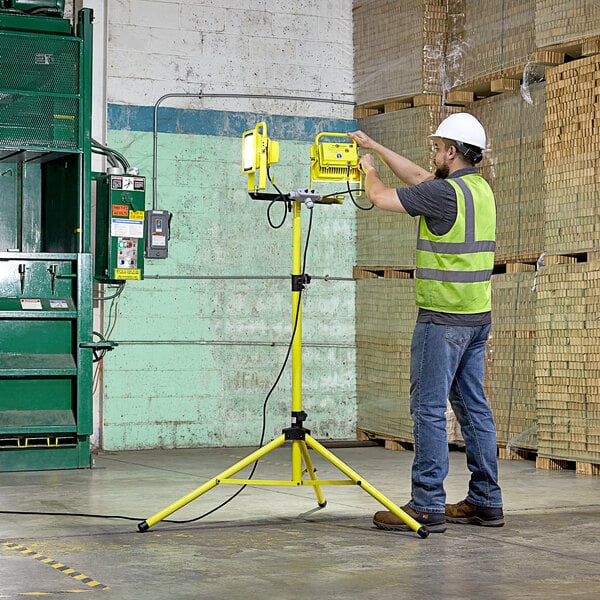 A man in a hard hat and safety vest adjusts a Lind Equipment LED dual head floodlight on a yellow tripod.
