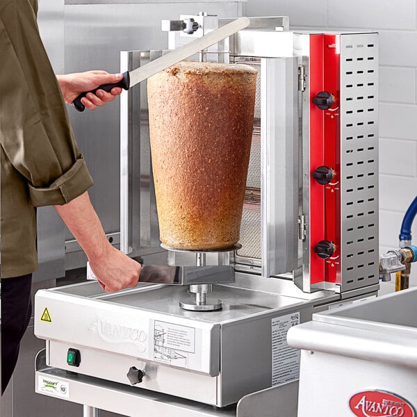 A person using an Avantco gas vertical broiler to cut gyro meat.