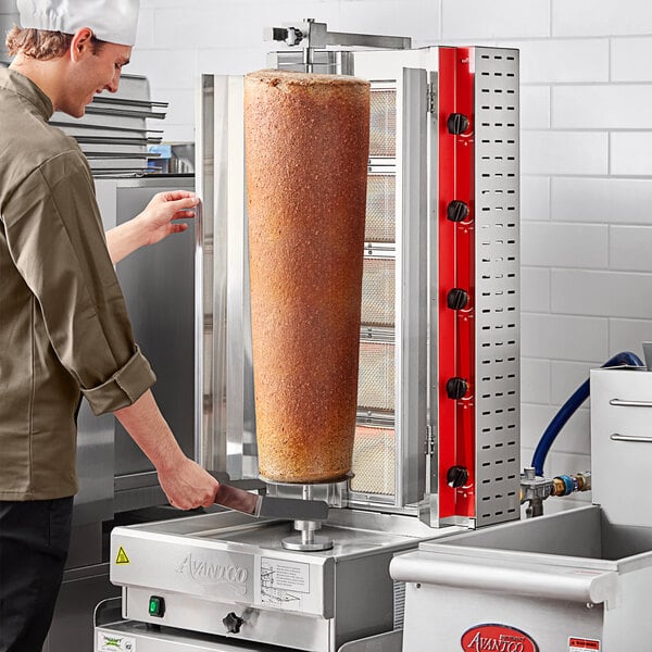 A man in a white hat using an Avantco gas vertical broiler to cook a large piece of meat.