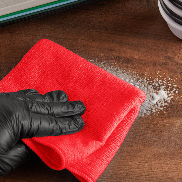 A hand in black gloves using a Lavex red microfiber cloth to wipe a table.