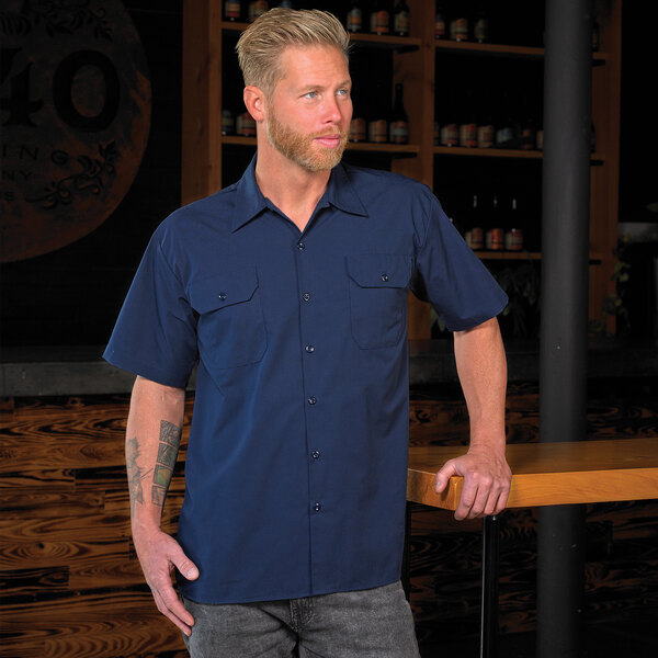 A man wearing a navy blue Mercer Culinary short sleeve work shirt standing next to a bar.