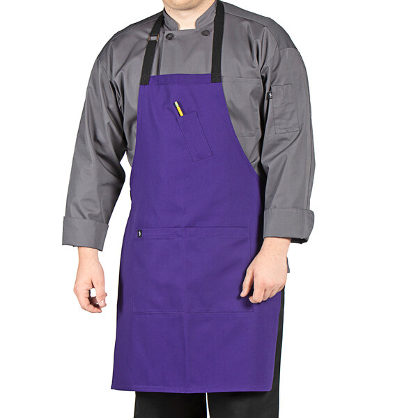 A man wearing a purple Uncommon Chef bib apron with black webbing standing in a professional kitchen.