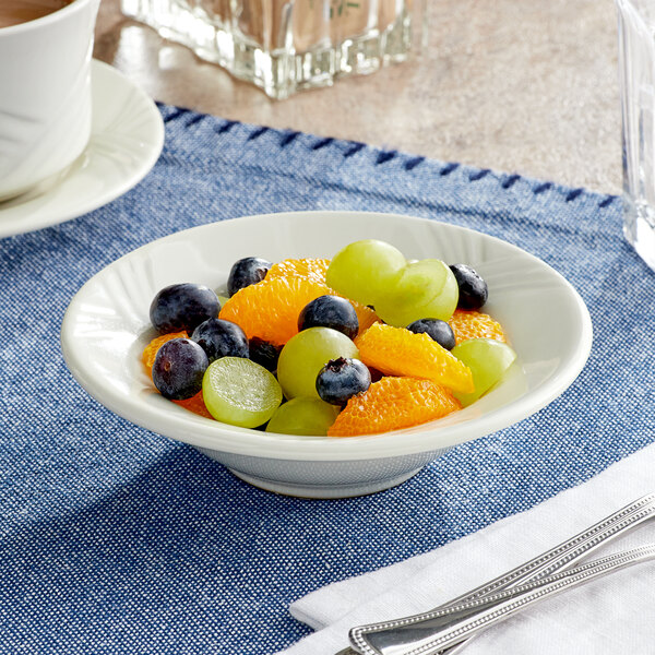 An Acopa ivory stoneware fruit bowl filled with oranges and grapes on a table with silverware.