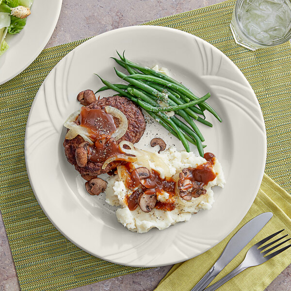 An Acopa Swell ivory stoneware plate with meat, mashed potatoes, and green beans on a table with a drink.