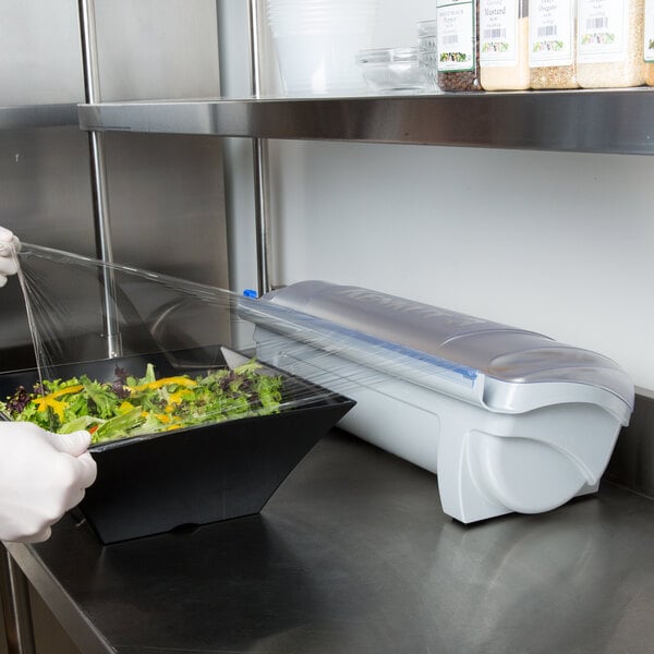 A person using a Tablecraft KenKut 3 plastic wrap dispenser to cover a salad in a white plastic container.