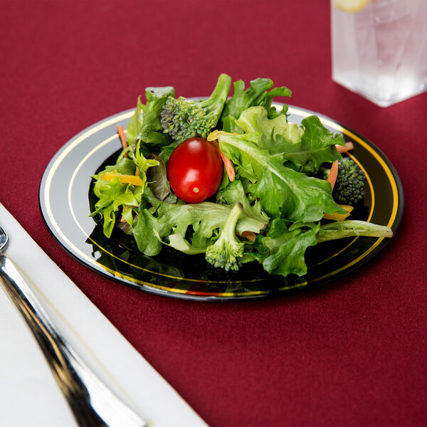 A Fineline Black plastic plate with gold bands holding salad with a cherry tomato and broccoli.