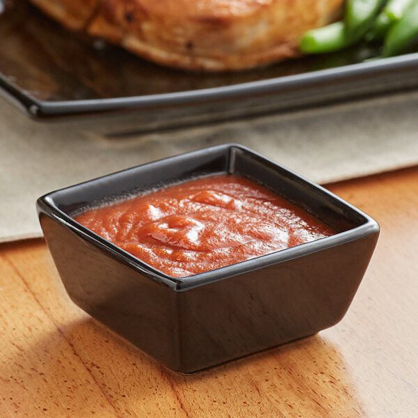 An Acopa glossy black square stoneware sauce cup filled with red sauce.
