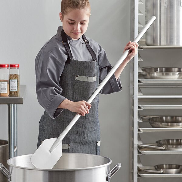 A woman in a chef's uniform using an American Metalcraft stainless steel paddle to stir a large pot.