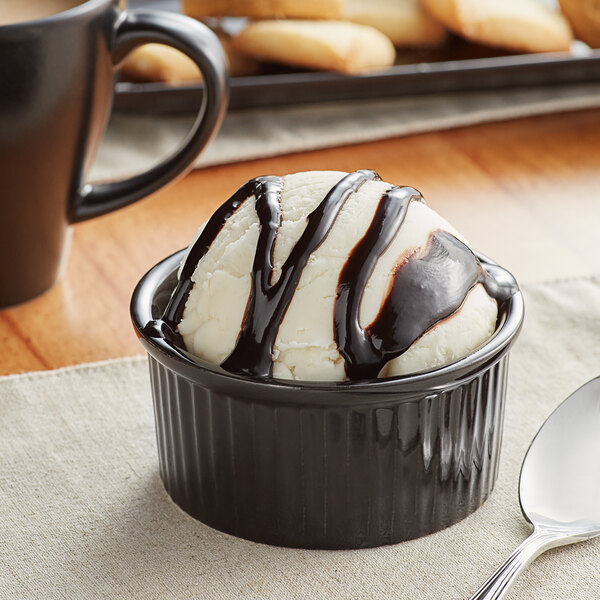 A glossy black fluted stoneware ramekin filled with a scoop of ice cream drizzled with chocolate sauce with a spoon next to it.