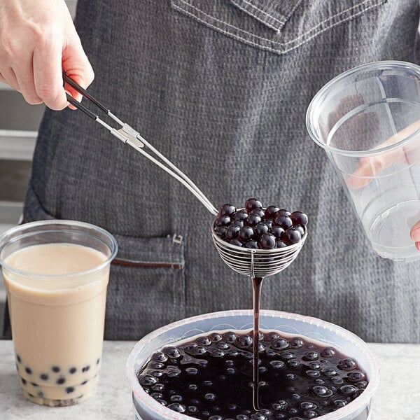 A person using a stainless steel wire scoop to pour blackberries into a clear plastic cup.