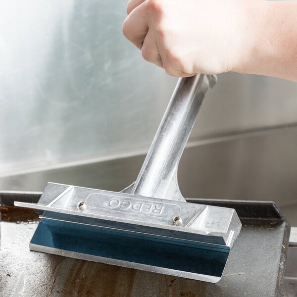 A hand using a Vollrath Redco Grill Scraper to clean a grill pan.