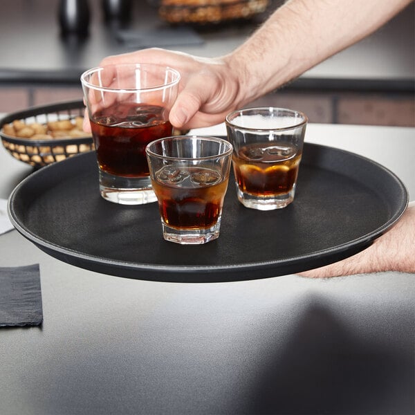 A person holding a Carlisle non-skid serving tray with three glasses of brown liquid.
