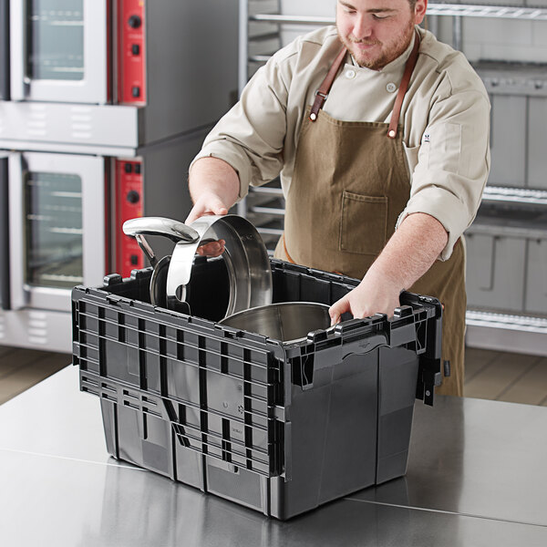 A man in a professional kitchen using a black Orbis Stack-N-Nest tote box with a hinged lid to store food.