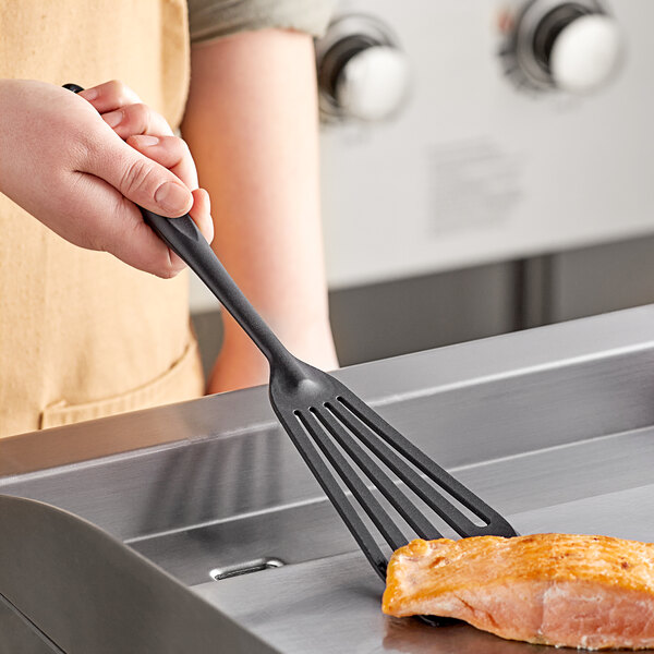 A person using a Tablecraft black silicone-coated stainless steel slotted fish/egg turner to flip salmon on a table.