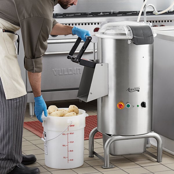 A man in a blue shirt and gloves using an Avantco 40 lb. floor potato peeler to peel a bucket of potatoes in a professional kitchen.