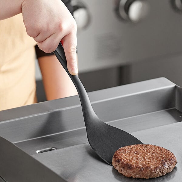 A hand holding a Tablecraft black silicone-coated stainless steel spatula over a burger.