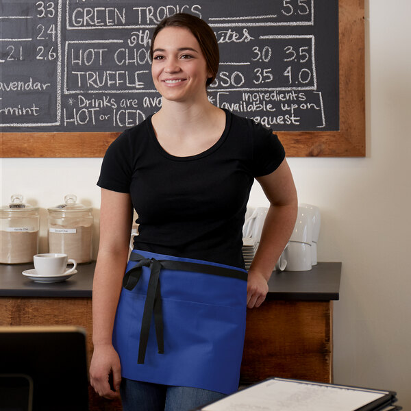 A woman wearing a Choice royal blue poly-cotton waist apron with black webbing standing in front of a chalkboard.