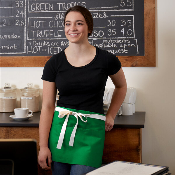 A woman wearing a Choice Kelly Green poly-cotton waist apron with natural webbing standing in front of a chalkboard.