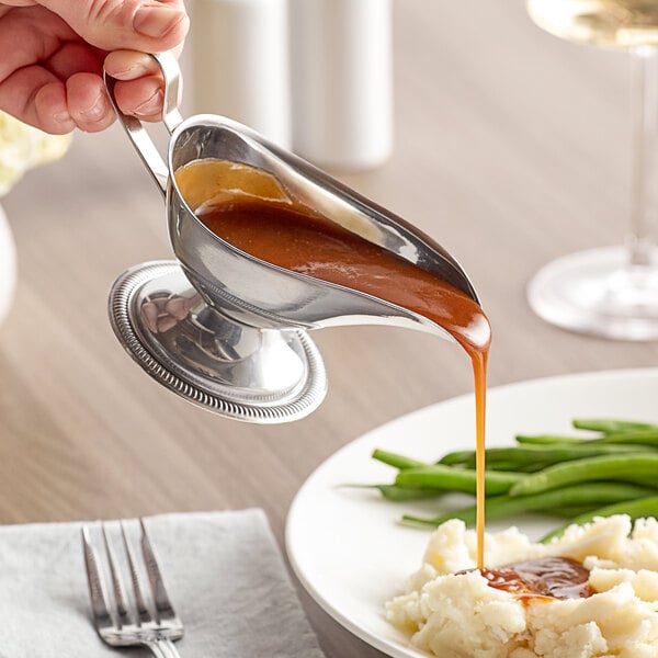 A person using a Vollrath stainless steel gravy boat to pour gravy over mashed potatoes.