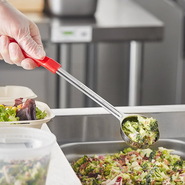 A person holding a Choice red solid portion spoon over a bowl of broccoli.