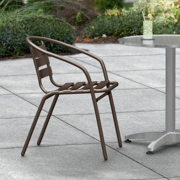 A brown Lancaster Table & Seating outdoor arm chair on a concrete patio next to a table with a glass of water.